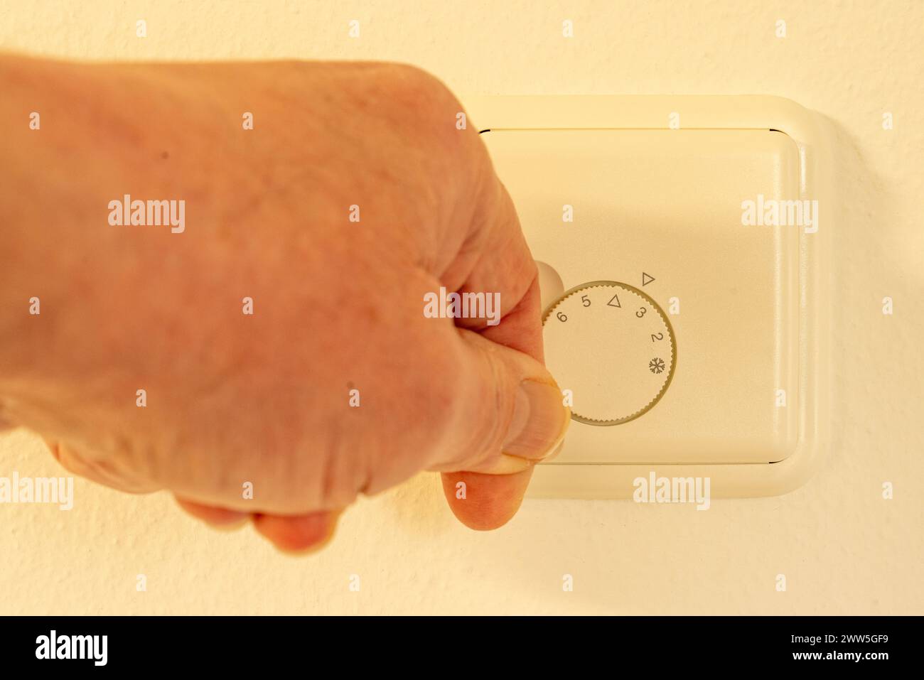 Hand changing the setting of a knob for regulating the room temperature on a white thermostat, mounted on a wall. Stock Photo