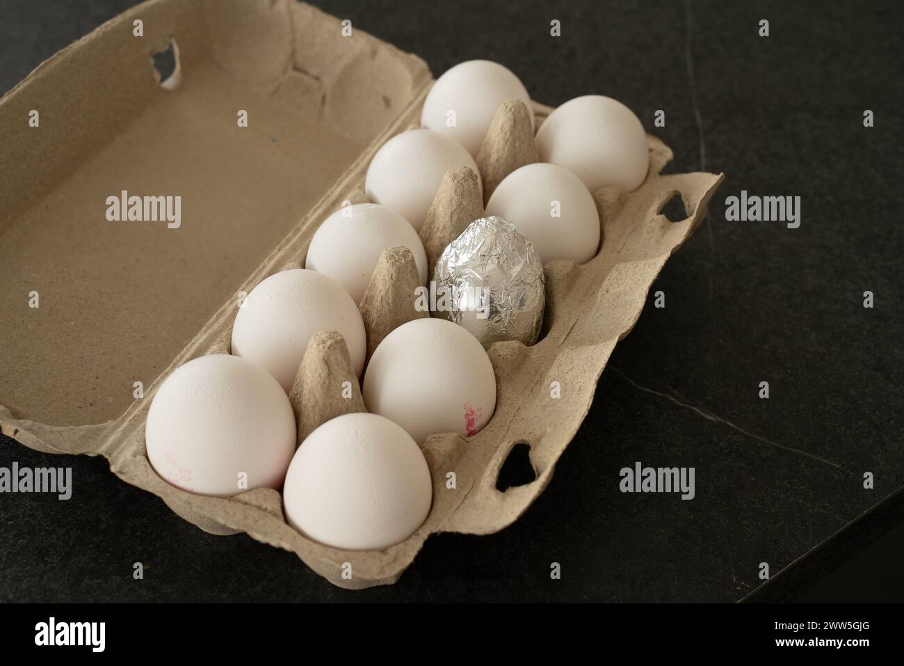 Brown open egg box on a dark table with ten white eggs in it. One egg in the middle is wrapped in silver foil, as easter egg. Stock Photo