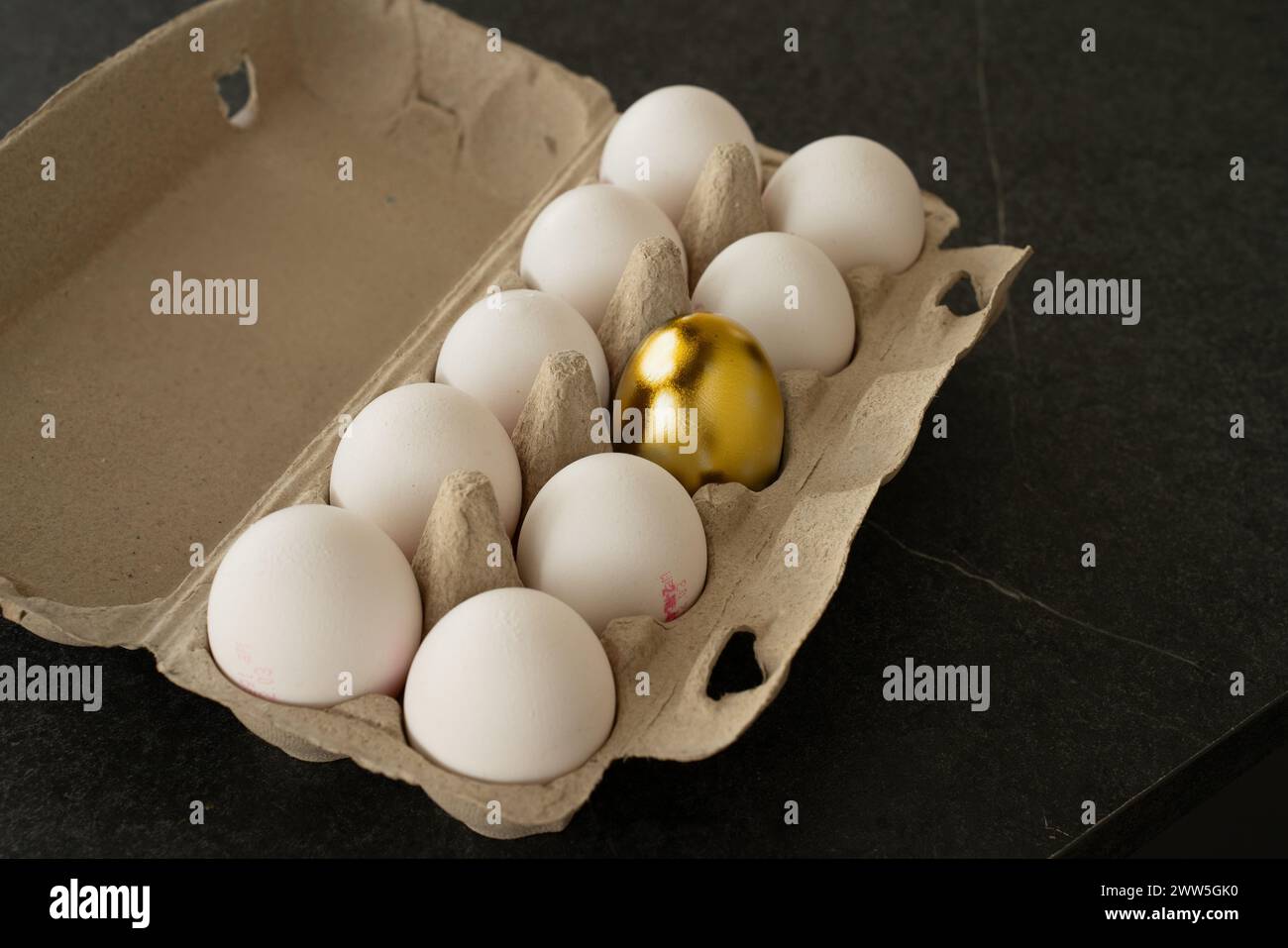 Brown open egg box on a dark table with ten white eggs in it. One egg in the middle is a golden easter egg. Stock Photo