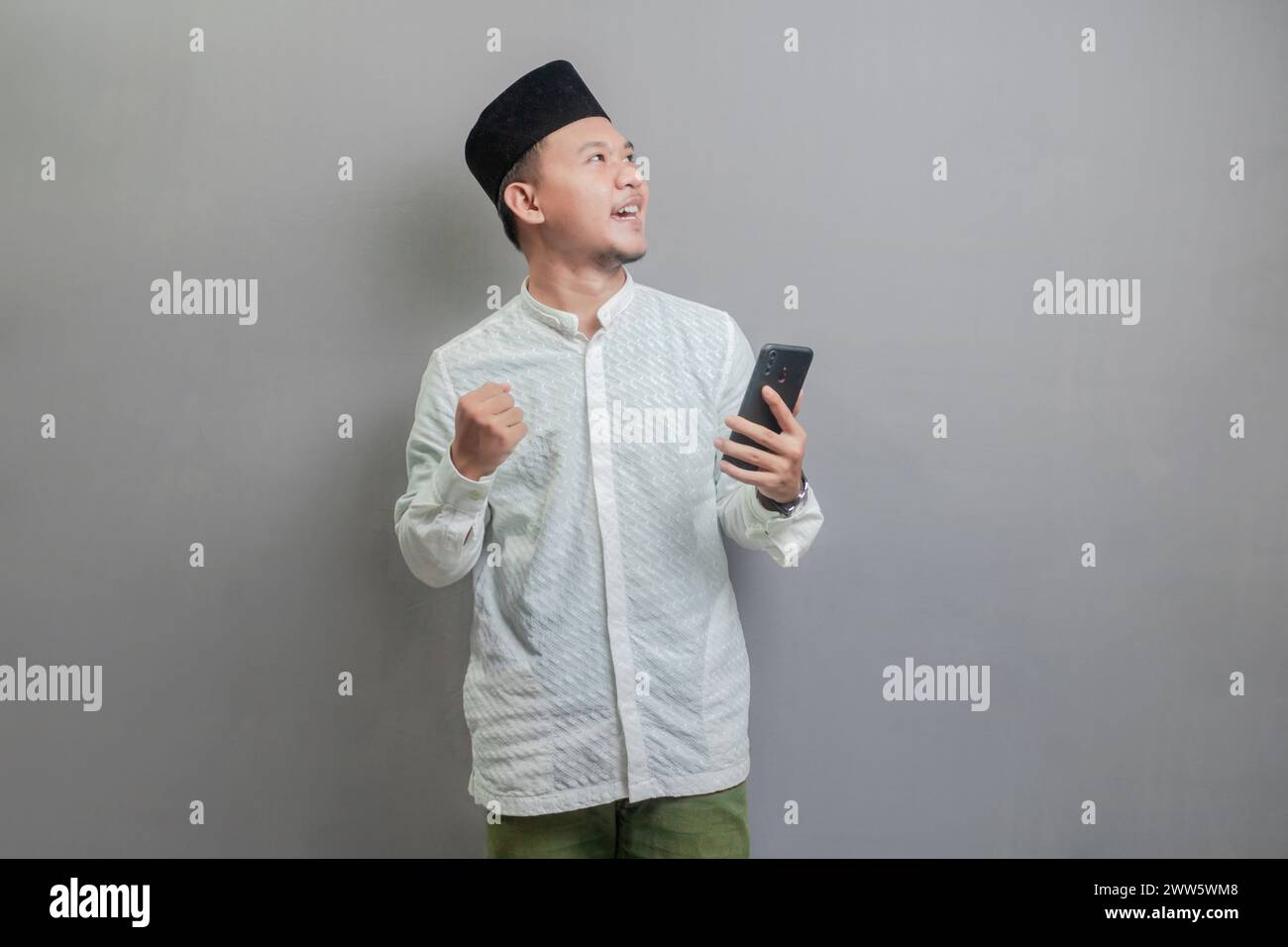 Asian Muslim man wearing a koko shirt and peci with shades of the fasting month, clenching his fist and holding his smartphone while looking at an emp Stock Photo