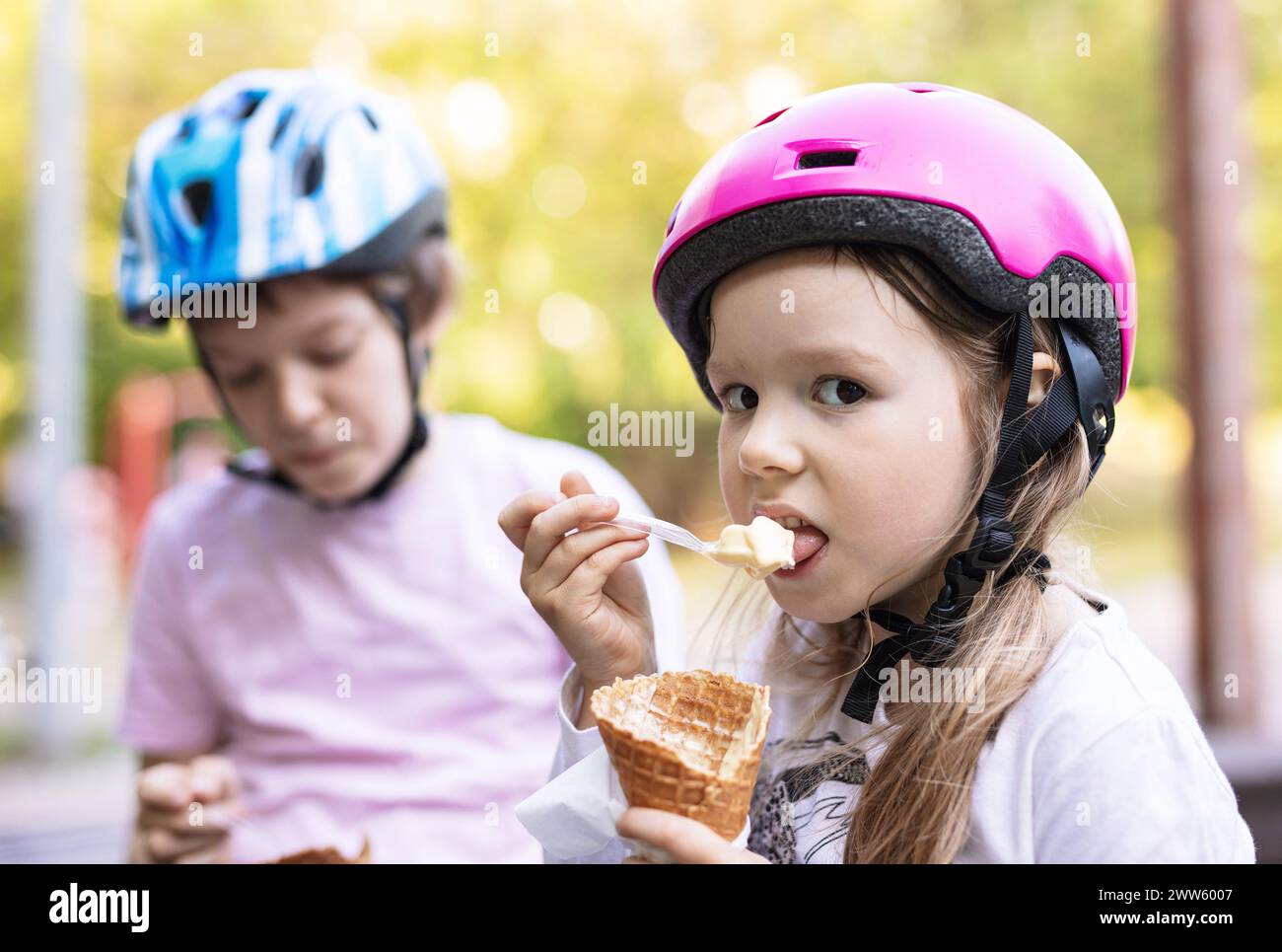 Young girl and boy eating ice cream and wearing helmets outdoors Stock Photo