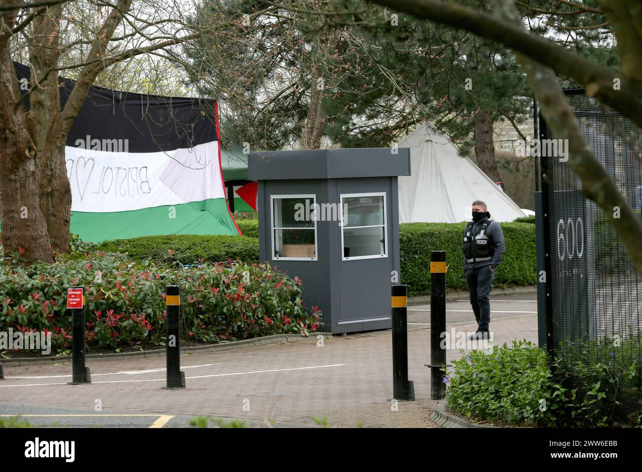 A security guard for Elbit Systems looks out onto the back of a huge Palestinian flag and a bell tent. Supporters of Palestine Action set up a protest camp outside Israeli arms company, Elbit Systems. The protest group have been targeting Elbit Systems at the eight businesses that operate in the UK. The protesters state 85% of drones flying in the Israeli defence force are provided by Elbit. These drones are oppressing Palestinians in Gaza and elsewhere and Palestine Action demand all Elbit Systems businesses in the UK close. As a result of persistent direct action Elbit has closed their offic Stock Photo