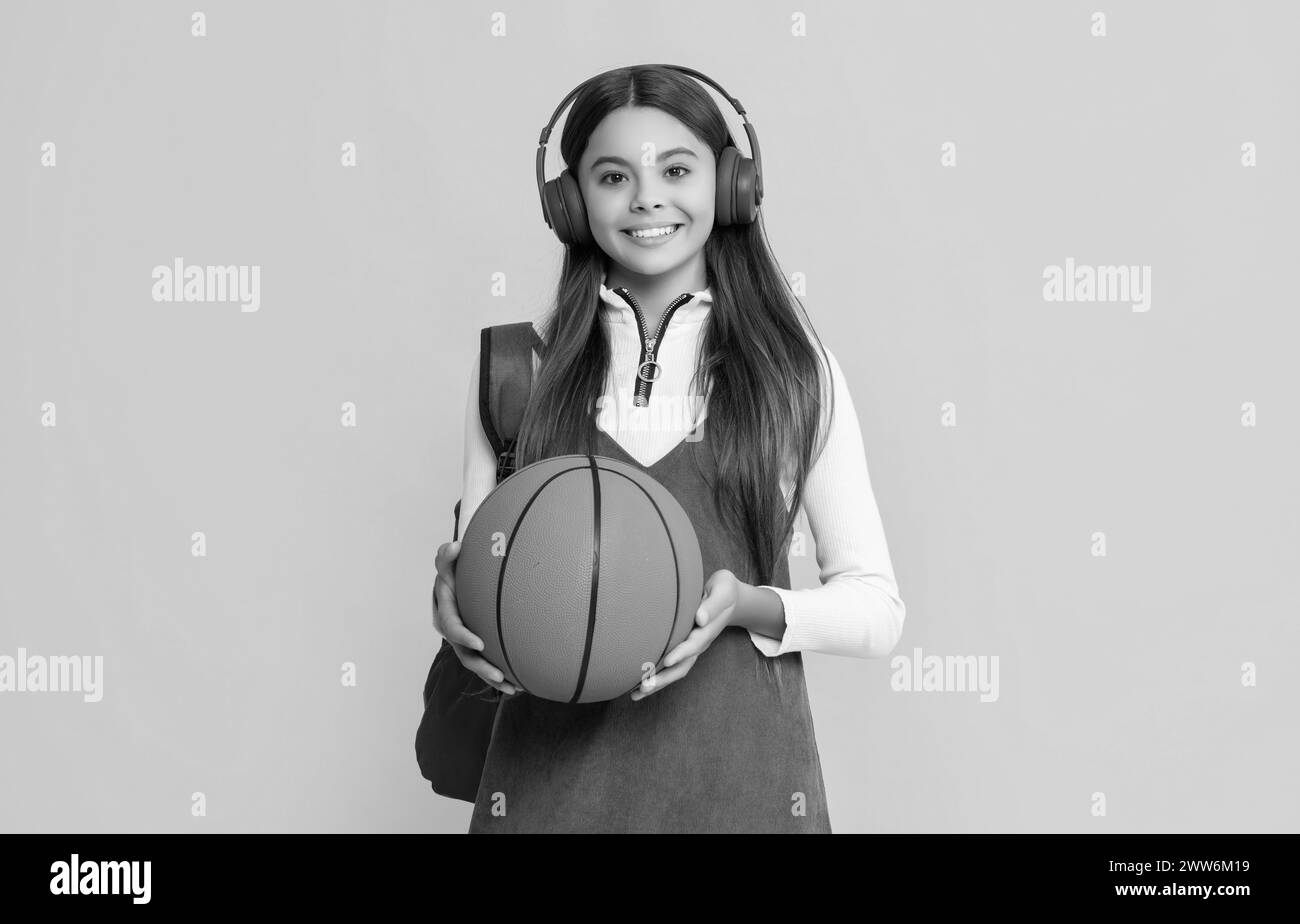 smiling kid in headphones with school backpack and basketball ball on yellow background Stock Photo