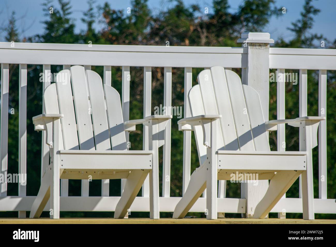 Two white Adirondack chairs on a deck with white railing and greenery ...