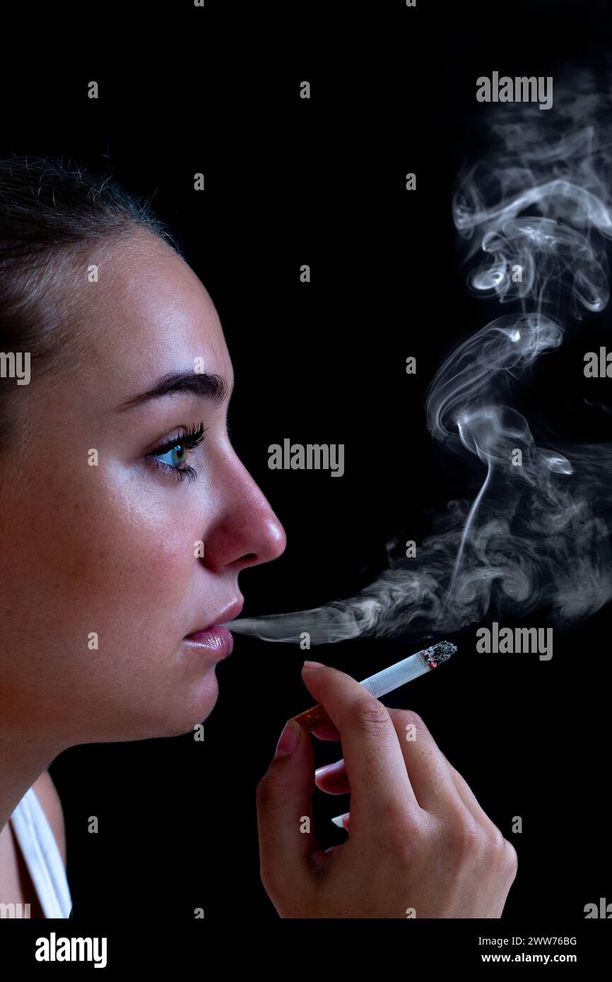 Close-up of a young woman smoking a cigarette against a black background, creating billows of smoke. Stock Photo