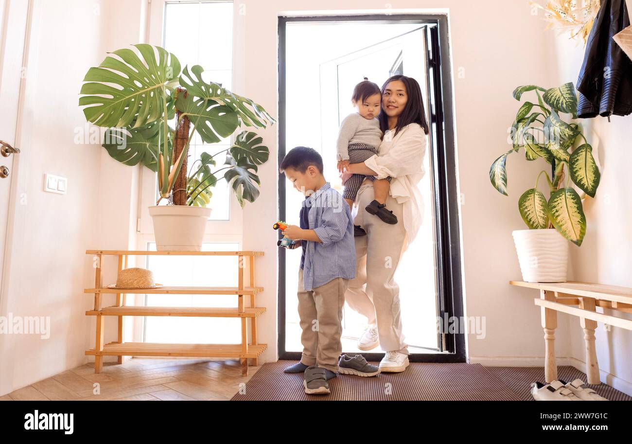 A happy Asian family. A young attractive woman helps to take off her little daughter shoes. A charming Korean mother and her children have returned ho Stock Photo