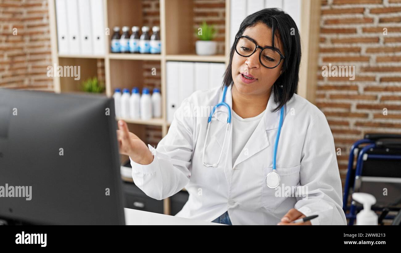 A professional hispanic woman in a white lab coat works diligently at her clinic computer amidst medical supplies. Stock Photo