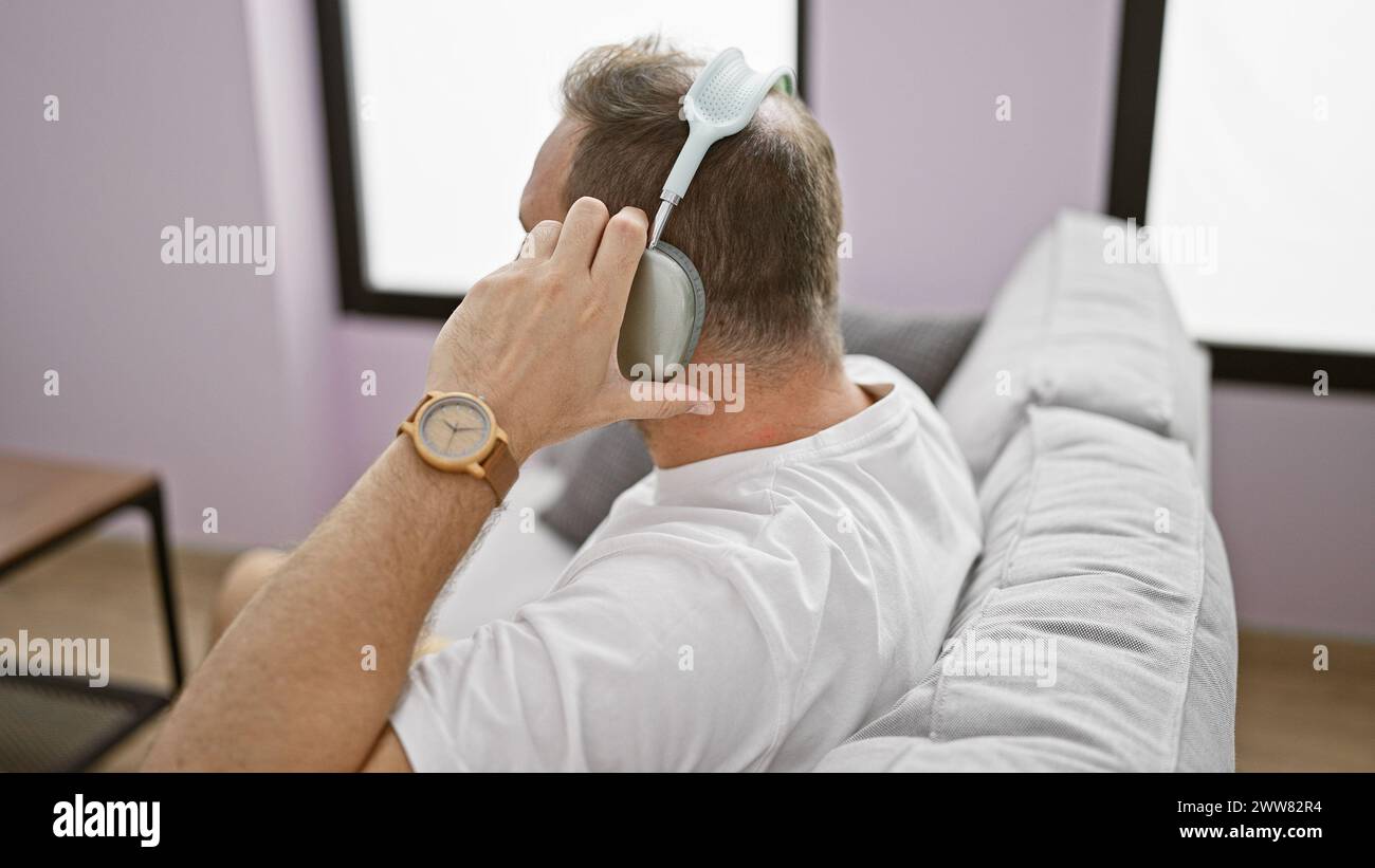 A middle-aged man with grey hair relaxes at home, massaging head with a handheld massager while sitting on a grey sofa. Stock Photo