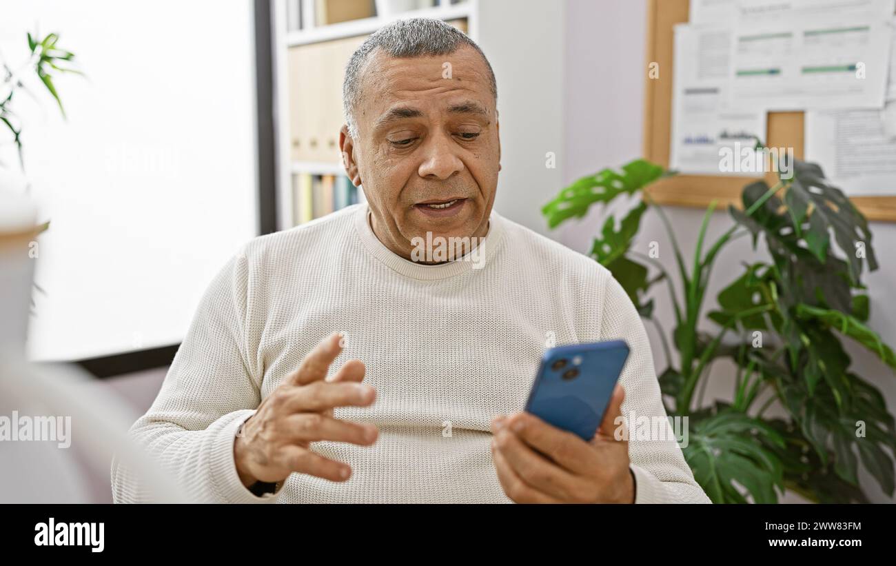 Mature hispanic man using smartphone in modern office setting, conveying connectivity and professionalism. Stock Photo