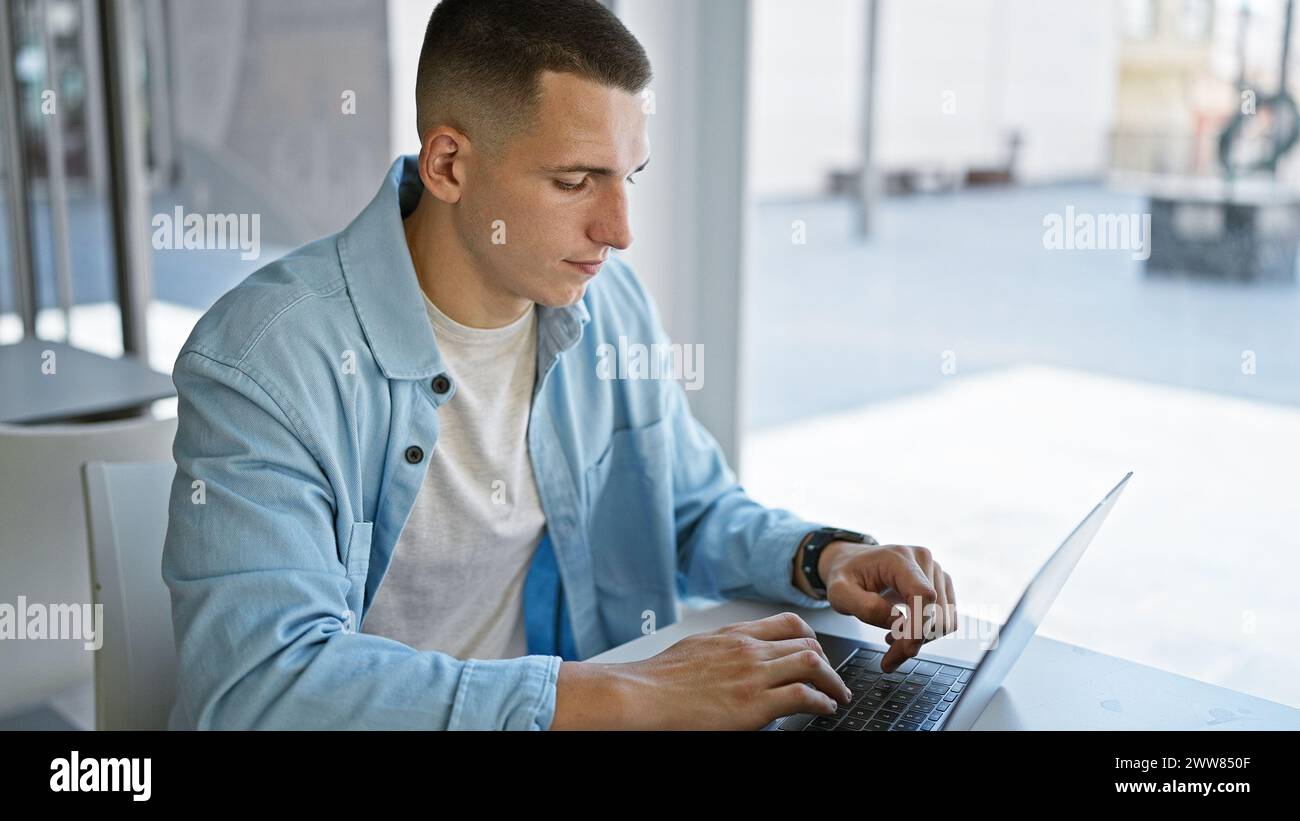 A focused young hispanic man using a laptop in a bright university library setting. Stock Photo
