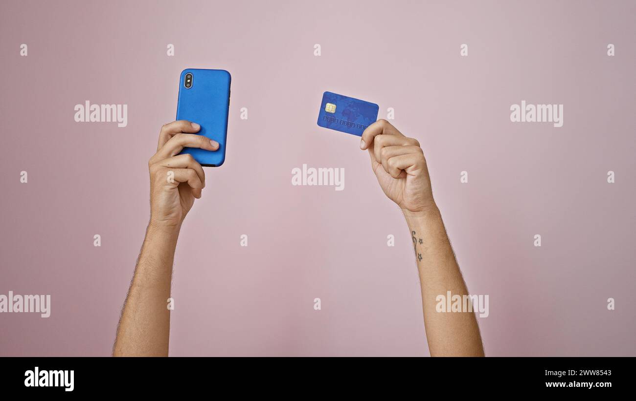 Man's tattooed arms holding smartphone and credit card against pink wall, depicting online payment or shopping. Stock Photo