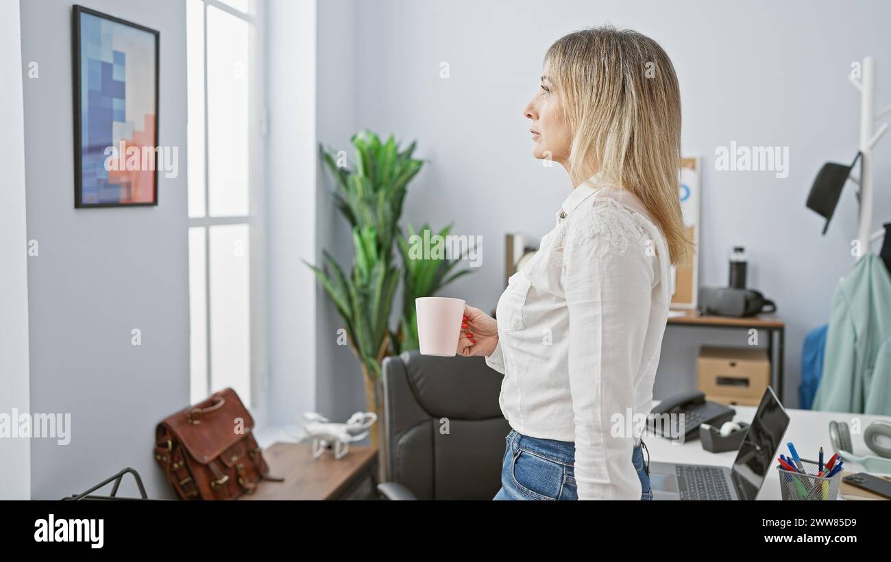 A middle-aged woman with blonde hair, standing thoughtfully in a modern office, holding a coffee cup. Stock Photo