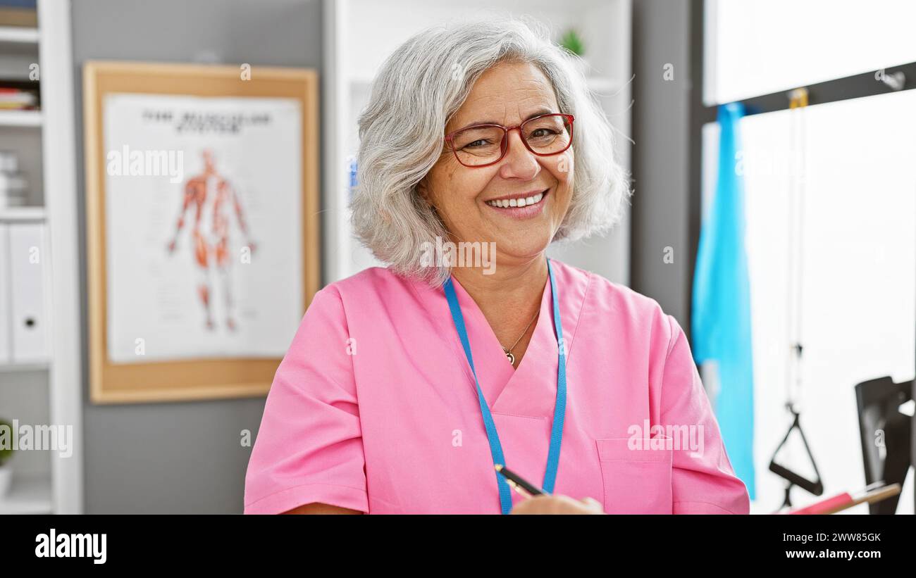Smiling middle-aged female healthcare professional in pink scrubs with glasses, standing in a clinic with an anatomy poster in the background. Stock Photo
