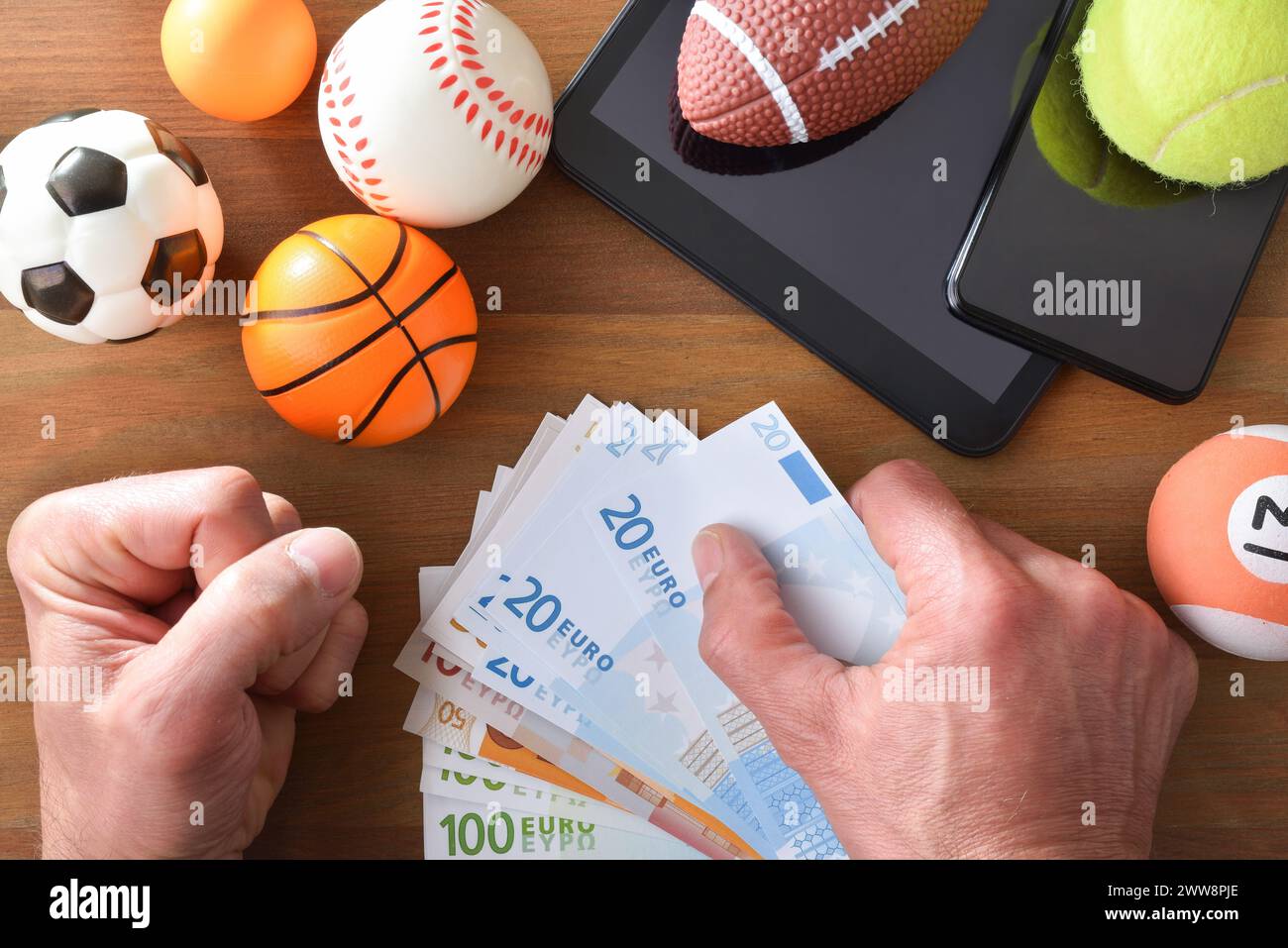 Concept of online sports betting with smartphone and tablet on wooden table with objects representing different sports and hands with money in hand to Stock Photo