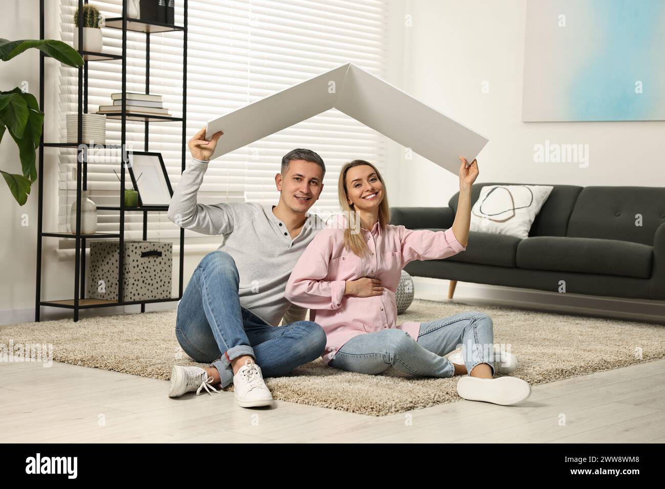 Young family housing concept. Pregnant woman with her husband sitting under cardboard roof on floor at home Stock Photo