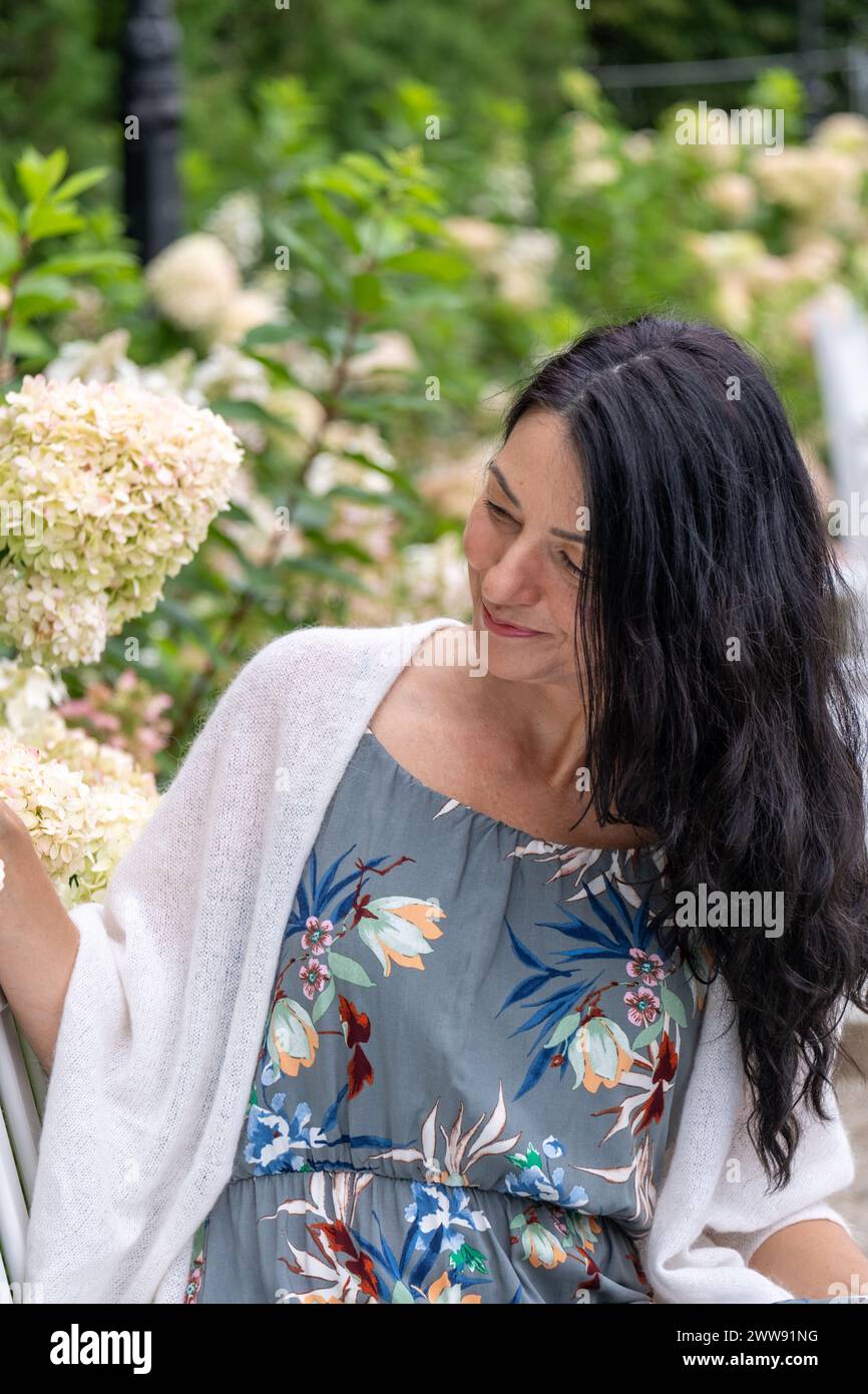 Adorned in a floral dress, a woman smiles gently amongst hydrangeas, her demeanor reflective of the wisdom and beauty found in life's natural cycles. Stock Photo