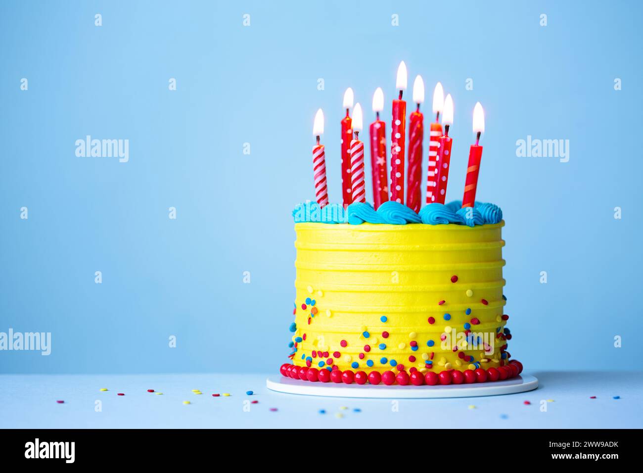 Colorful celebration birthday cake with yellow frosting and red birthday candles against a blue background Stock Photo