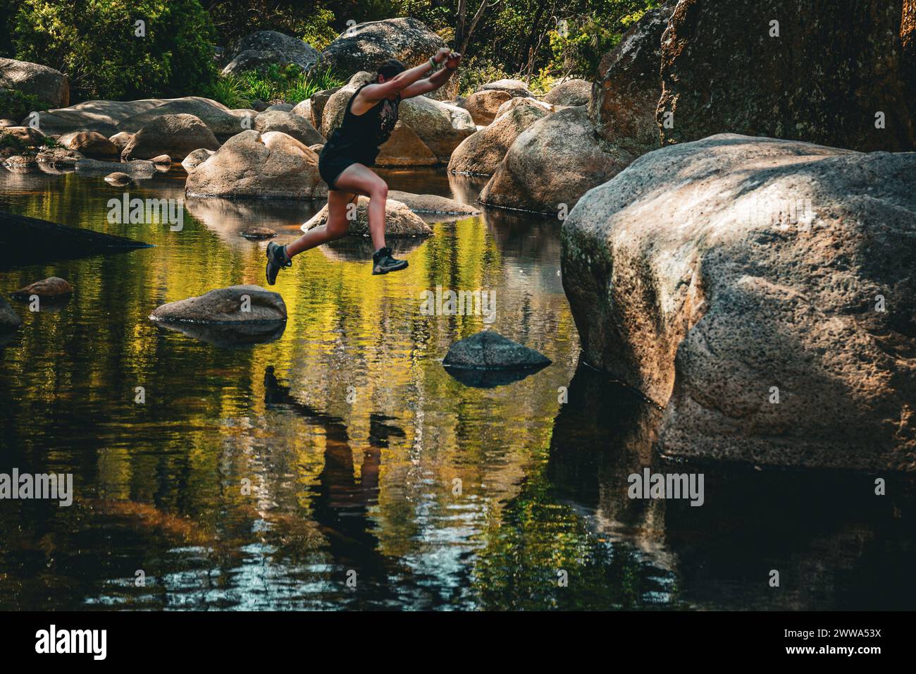 Hiker dynamically jumping across river stones surrounded by serene nature. Stock Photo