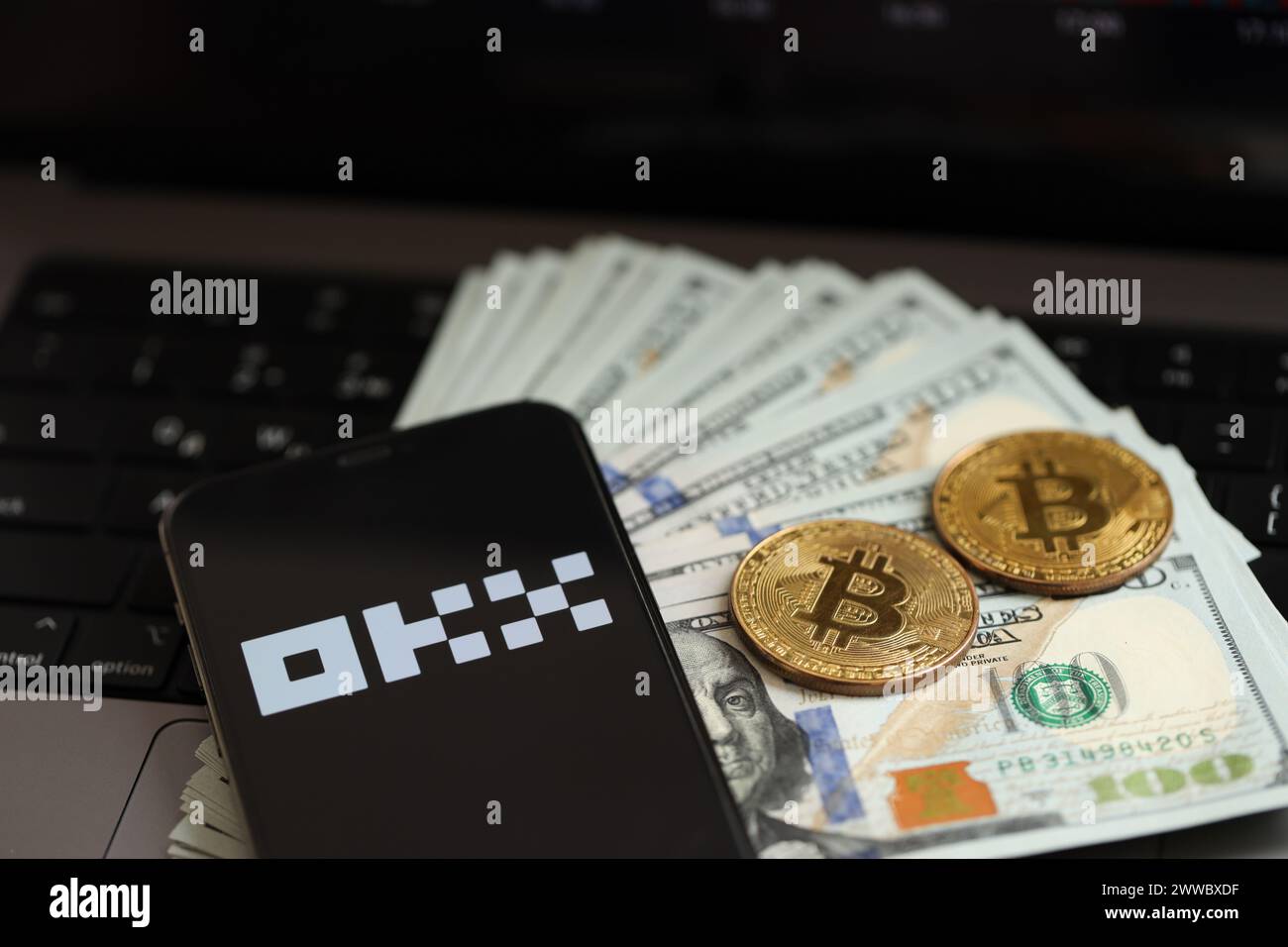 KYIV, UKRAINE - MARCH 15, 2024 OKX logo on iPhone display screen with dollars and bitcoins on MacBook. Cryptocurrency exchange portal Stock Photo