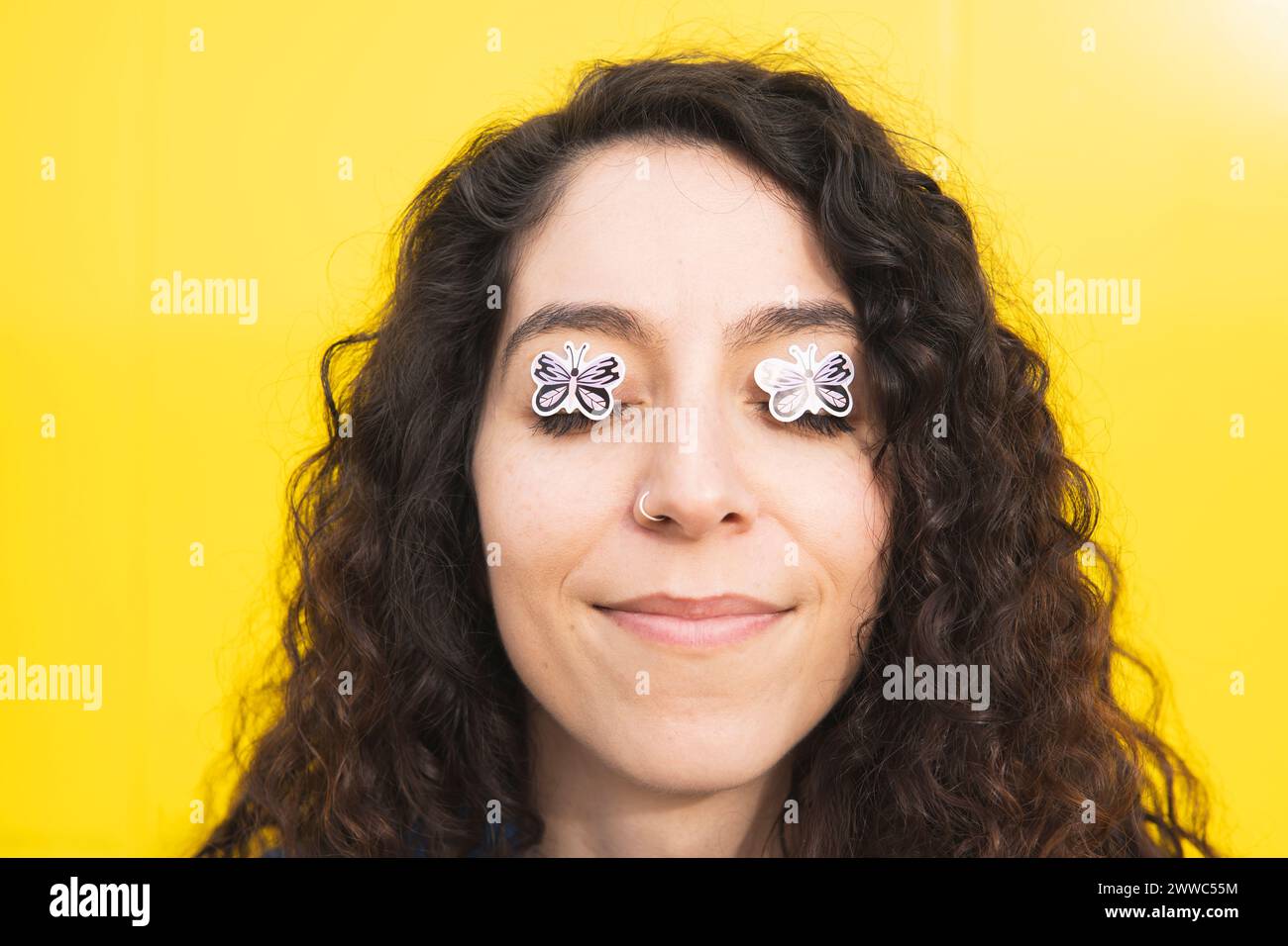 Smiling curly haired woman with butterfly stickers on eyes against yellow background Stock Photo