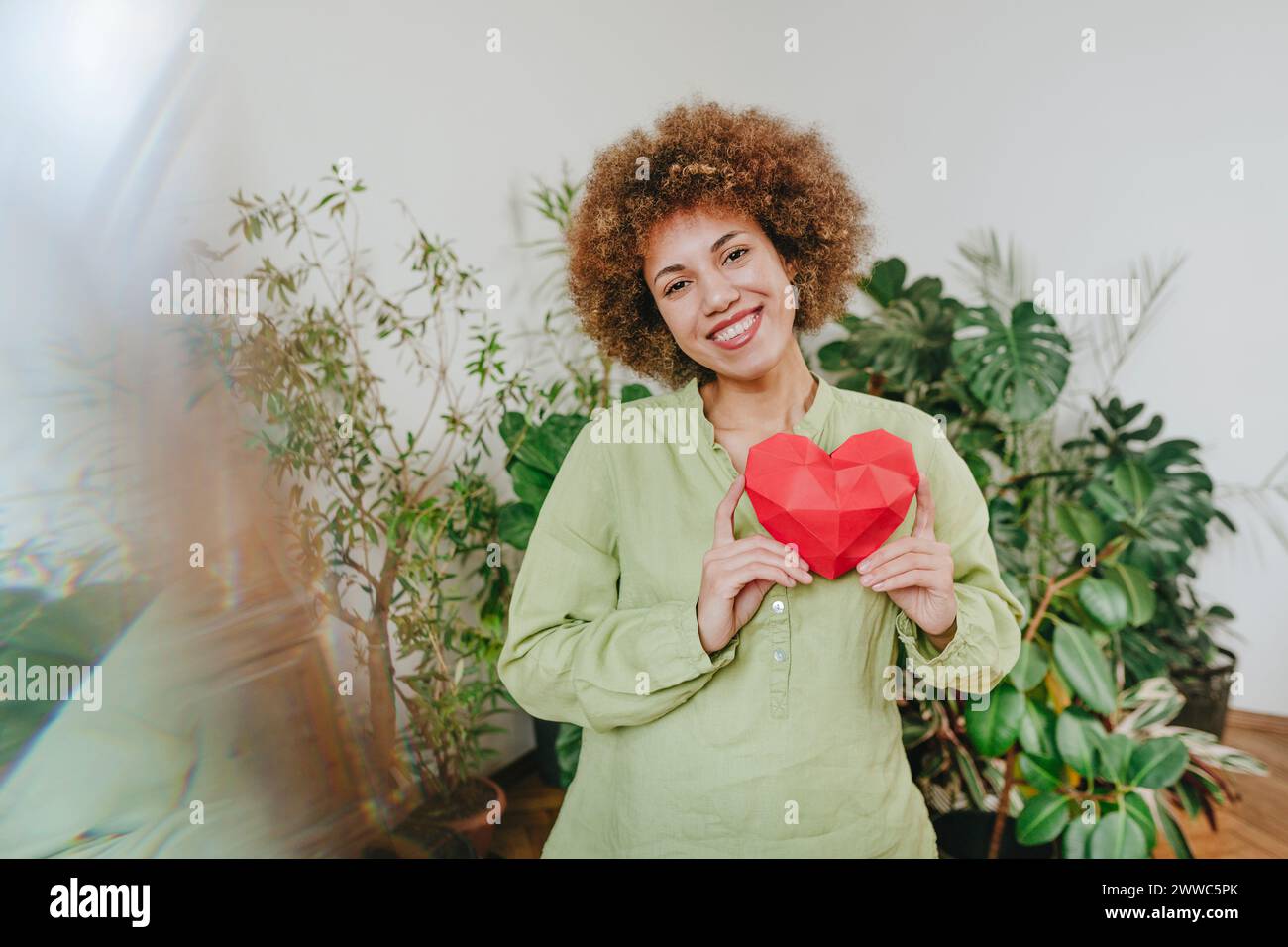 Happy young woman holding red paper heart near plants Stock Photo