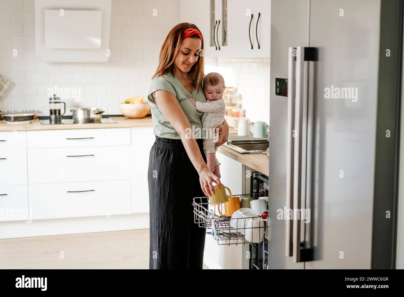 Woman carrying baby and taking out cup from dishwasher Stock Photo