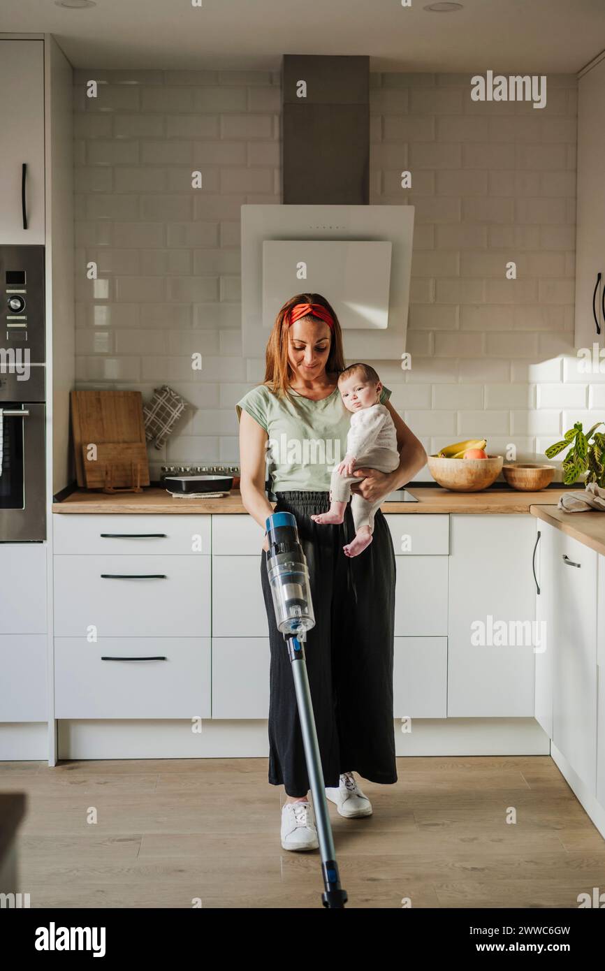 Woman carrying baby daughter and cleaning with vacuum cleaner at home Stock Photo