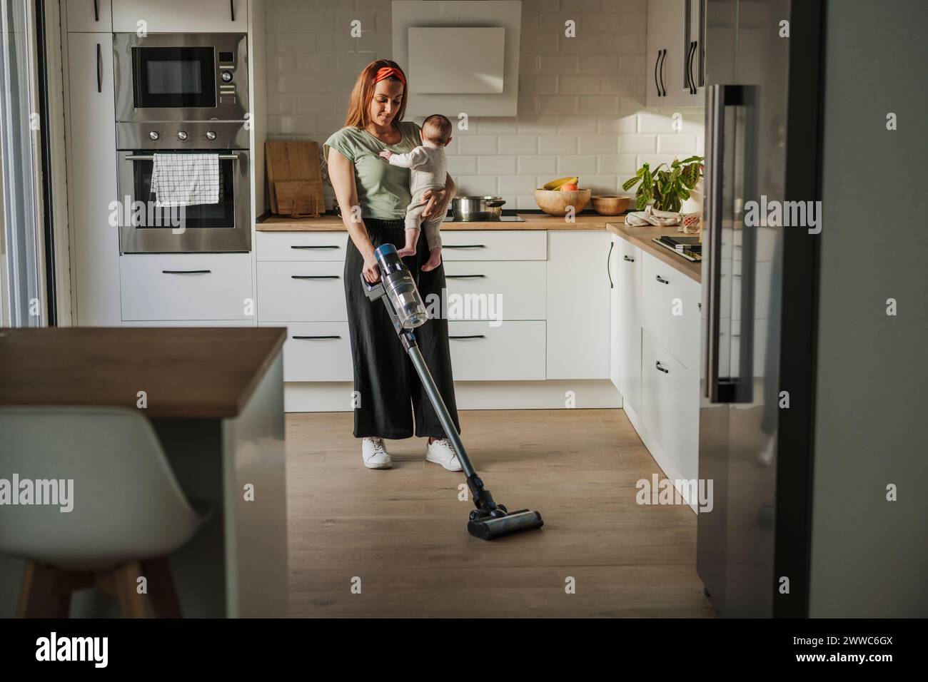Woman carrying baby daughter and cleaning kitchen with vacuum cleaner Stock Photo