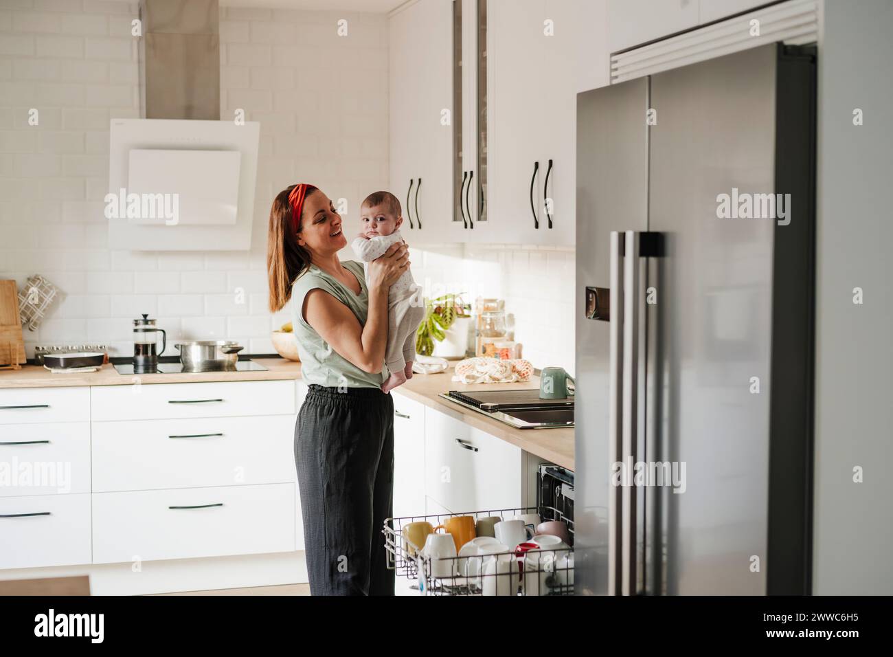 Happy woman carrying baby daughter in kitchen at home Stock Photo