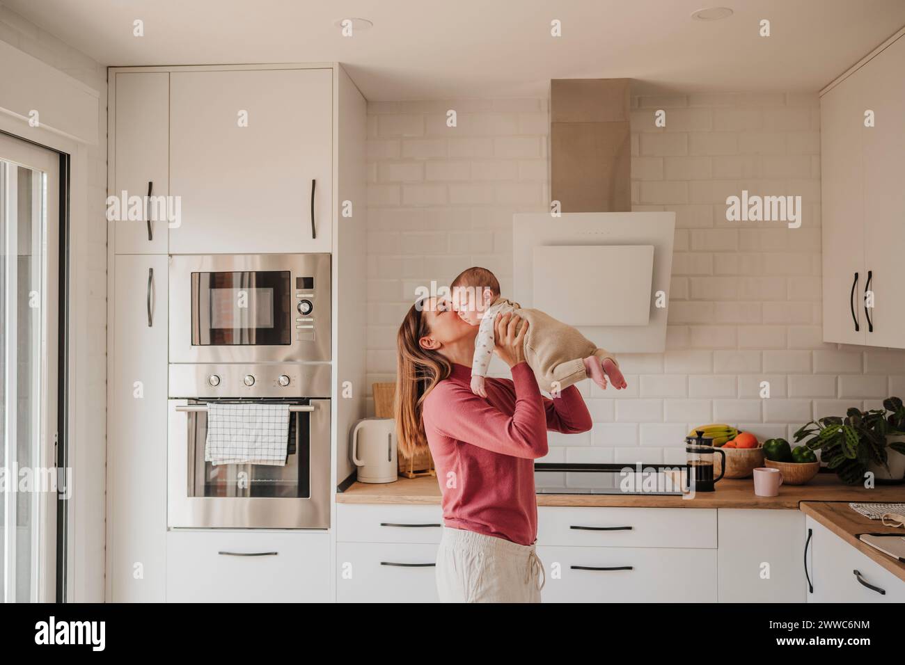 Mother kissing baby daughter in kitchen at home Stock Photo