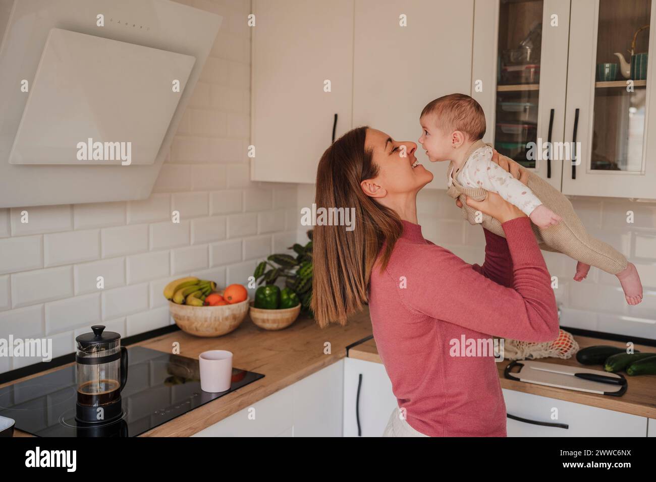 Smiling mother embracing and playing with baby daughter in kitchen Stock Photo