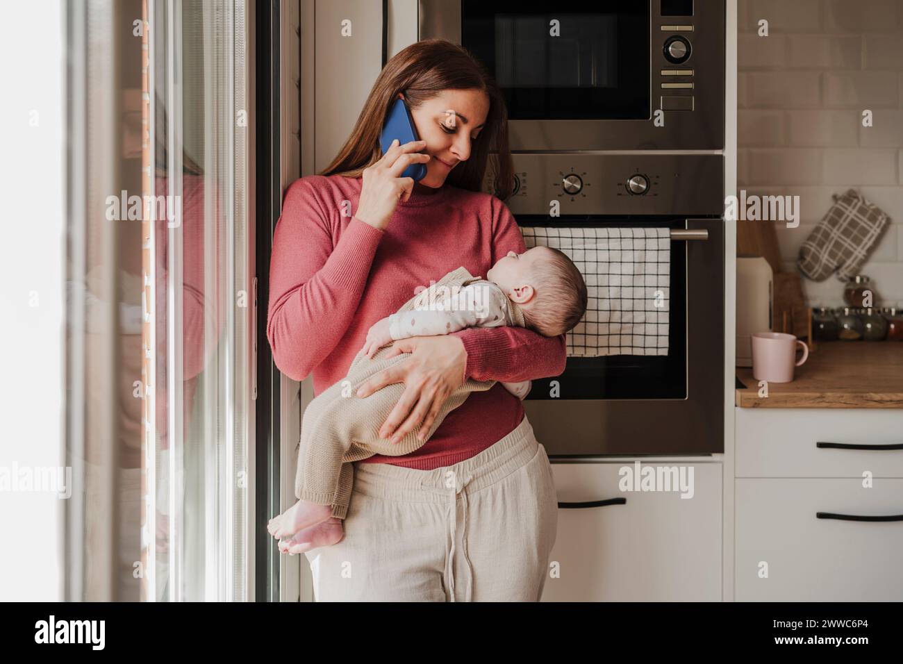 Smiling woman talking on smart phone and carrying baby daughter in kitchen Stock Photo