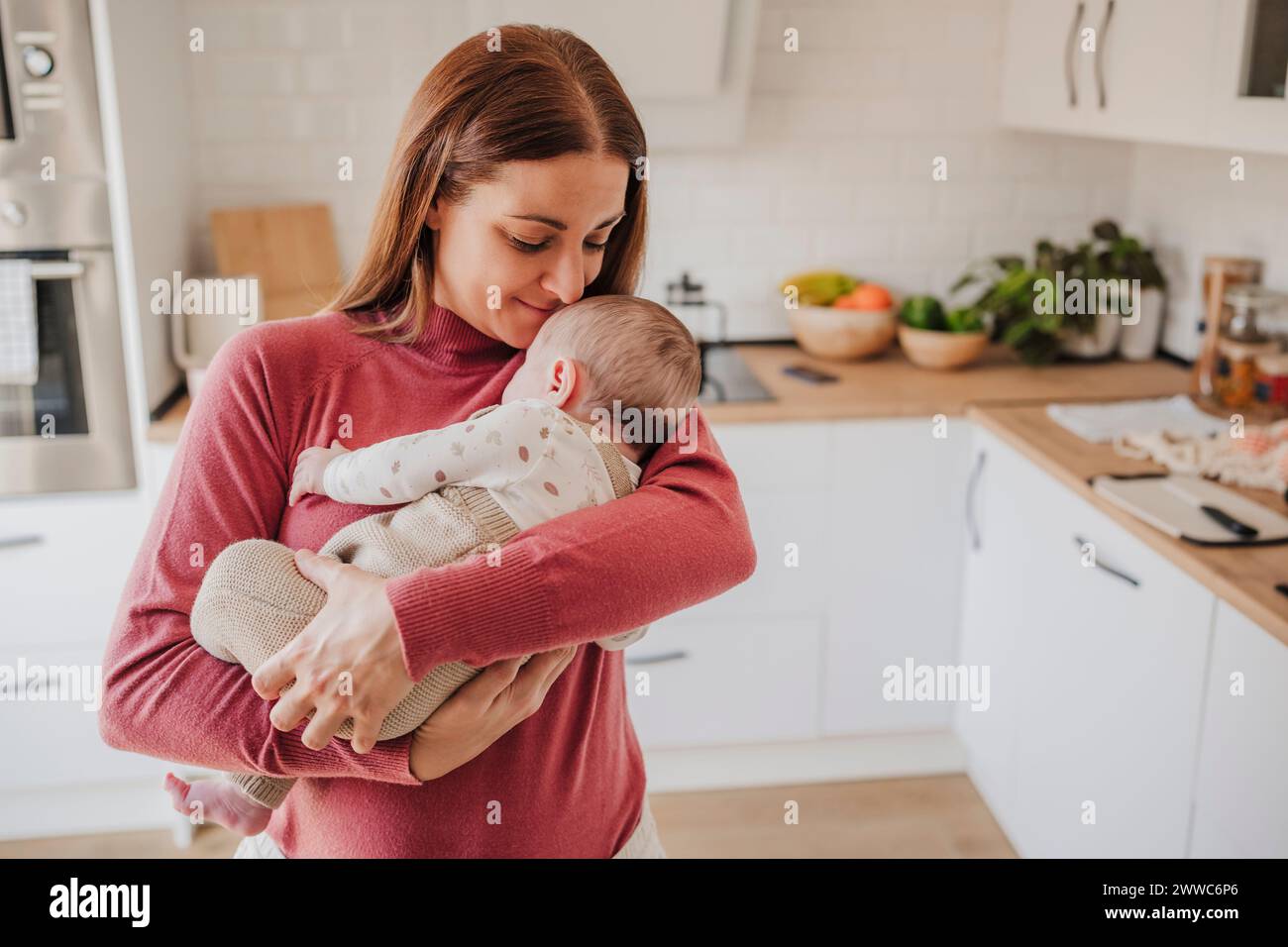 Smiling woman embracing baby daughter in kitchen at home Stock Photo