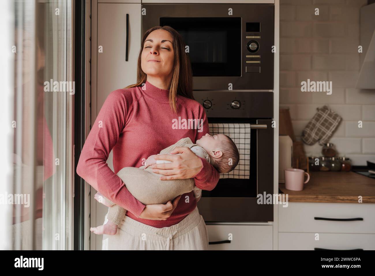 Smiling woman standing with eyes closed and carrying baby girl at home Stock Photo