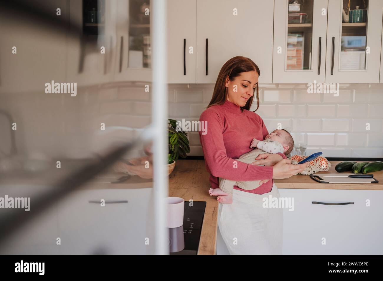 Woman carrying sleeping baby daughter and using smart phone in kitchen Stock Photo