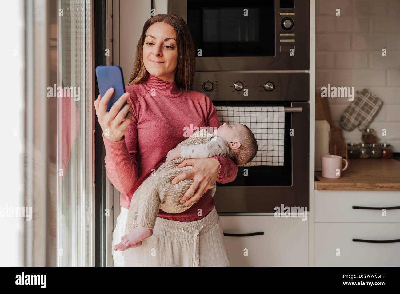 Smiling woman carrying baby girl and using smart phone in kitchen Stock Photo