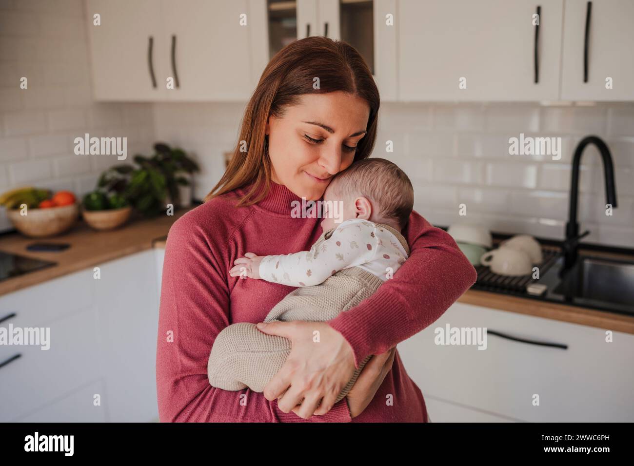 Smiling woman embracing baby daughter in kitchen Stock Photo