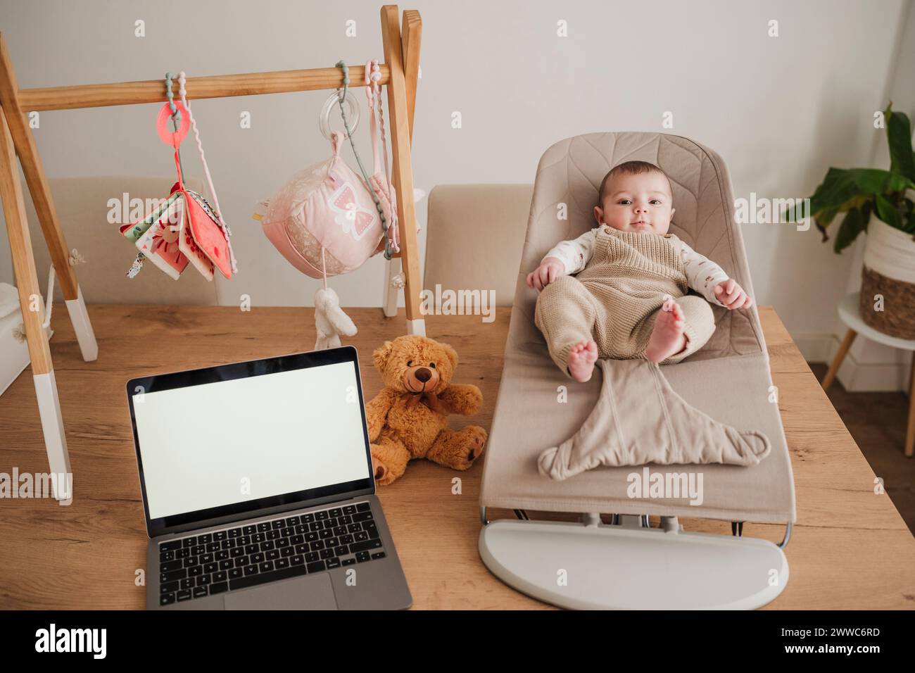 Cute baby girl sitting on bouncer chair near laptop at home Stock Photo