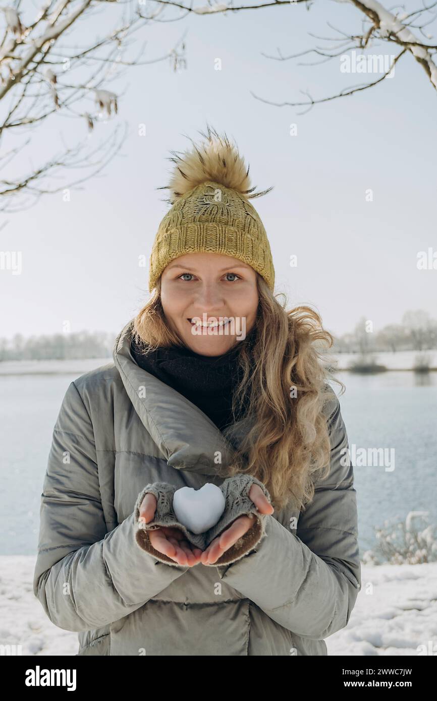 Happy woman holding heart shaped snowball near lake Stock Photo