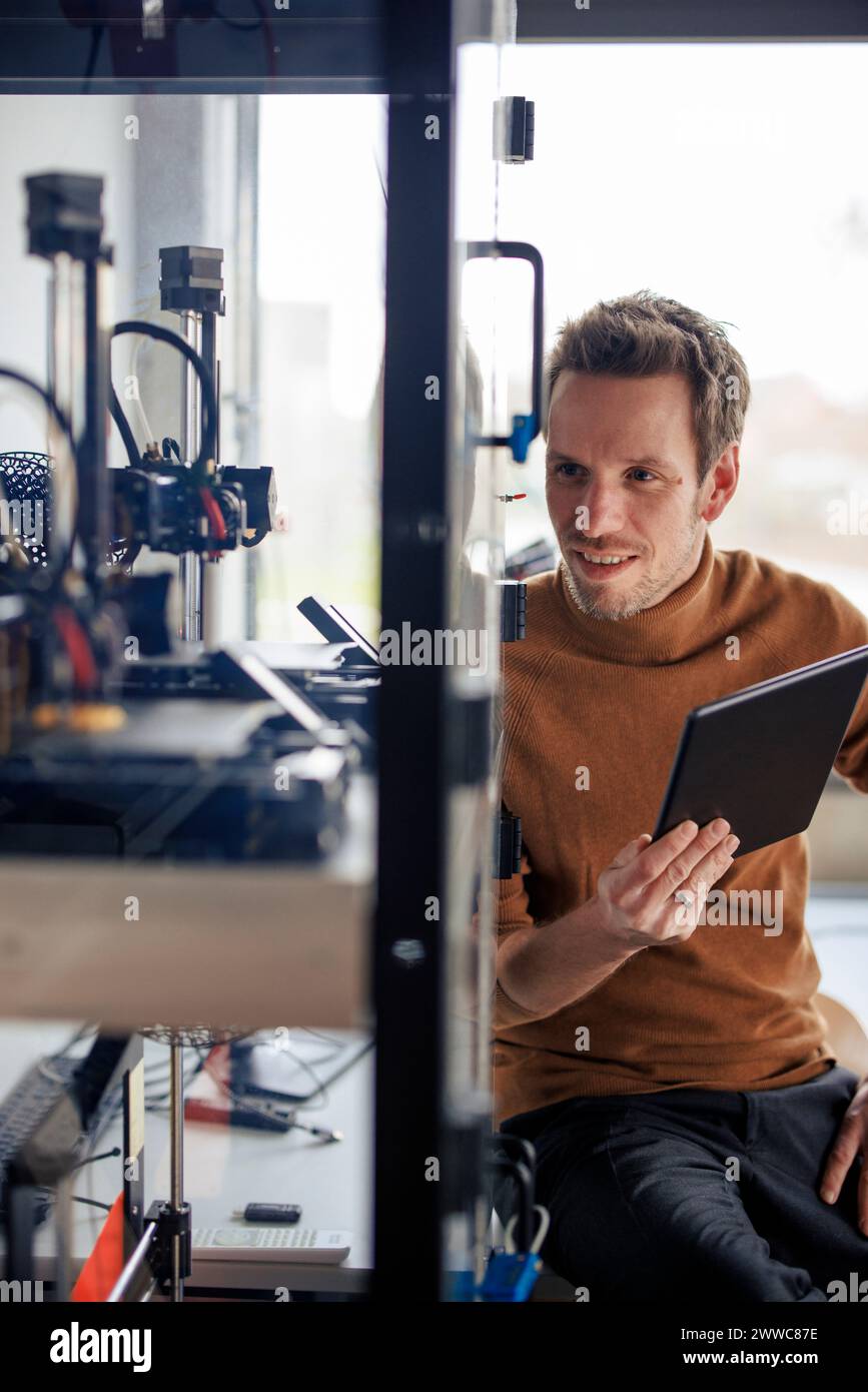 Smiling engineer holding tablet PC and looking at 3d printing machine in workshop Stock Photo