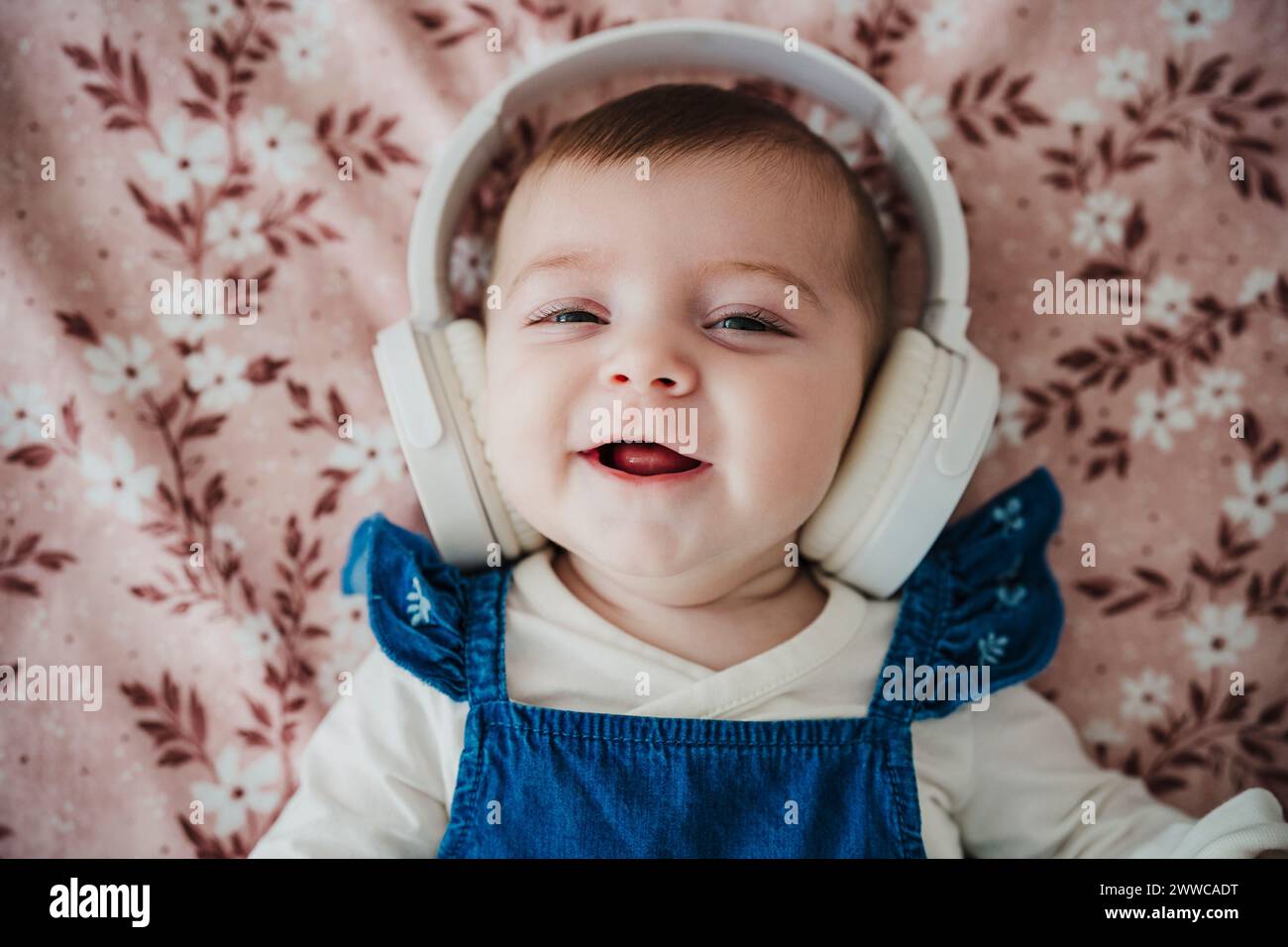 Cute baby girl wearing wireless headphones lying on bed Stock Photo