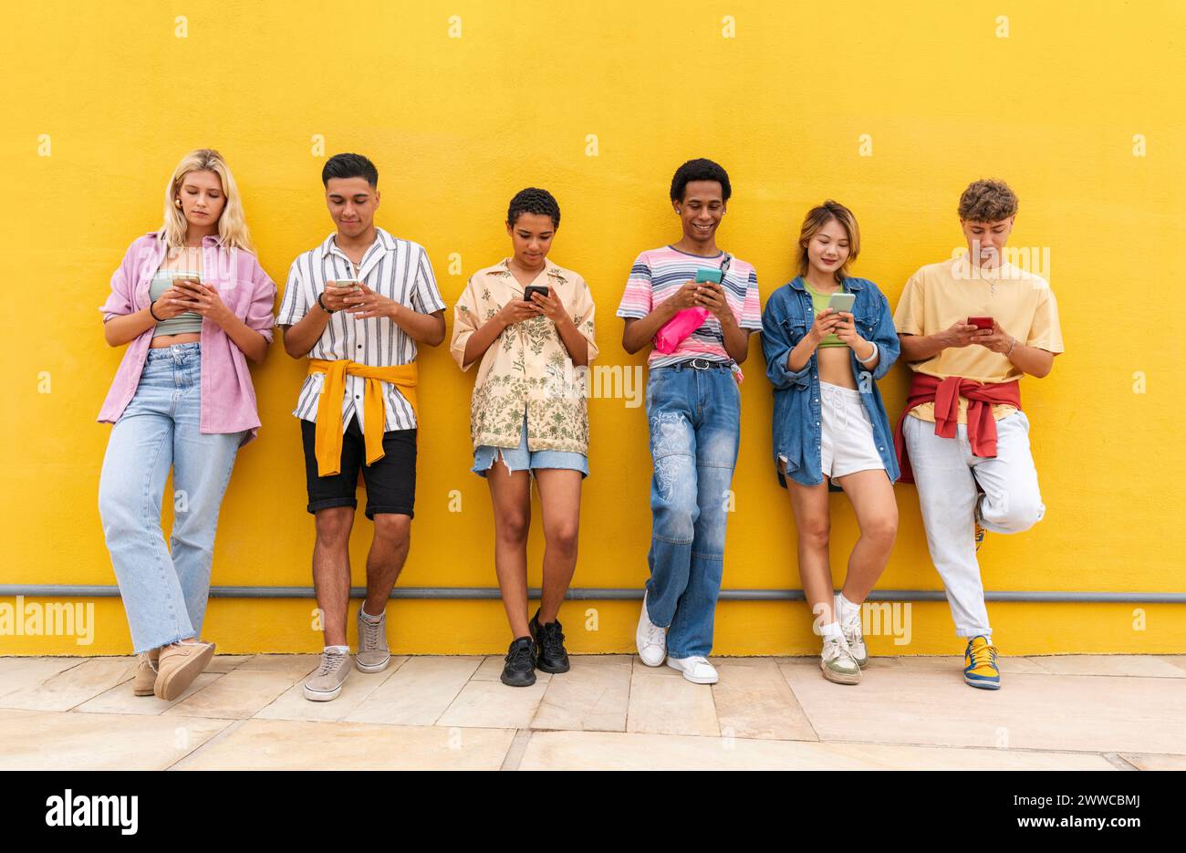 Group of friends leaning against yellow wall using their smartphones Stock Photo