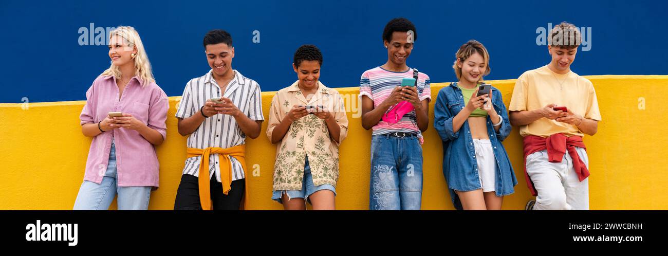 Group of friends leaning against yellow wall using their smartphones Stock Photo