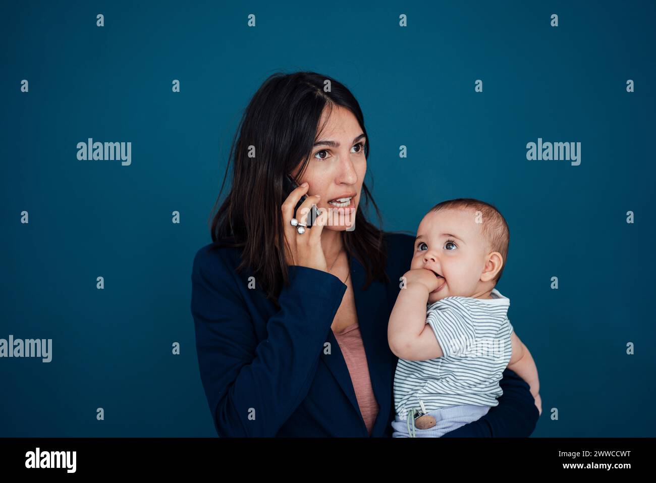 Busy businesswoman carrying daughter talking on mobile phone in front of blue background Stock Photo