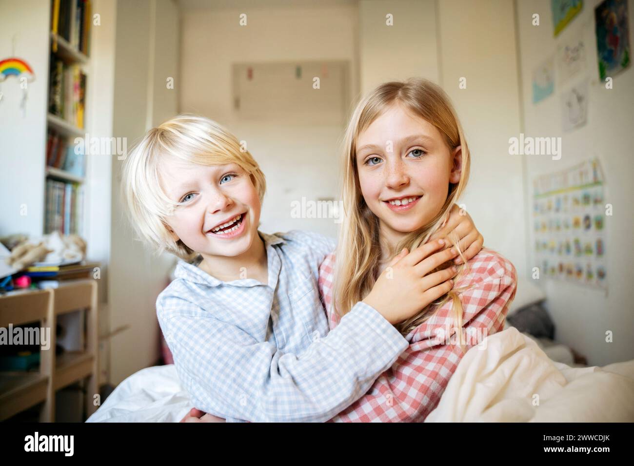 Smiling boy sitting with arm around sister in bedroom at home Stock Photo