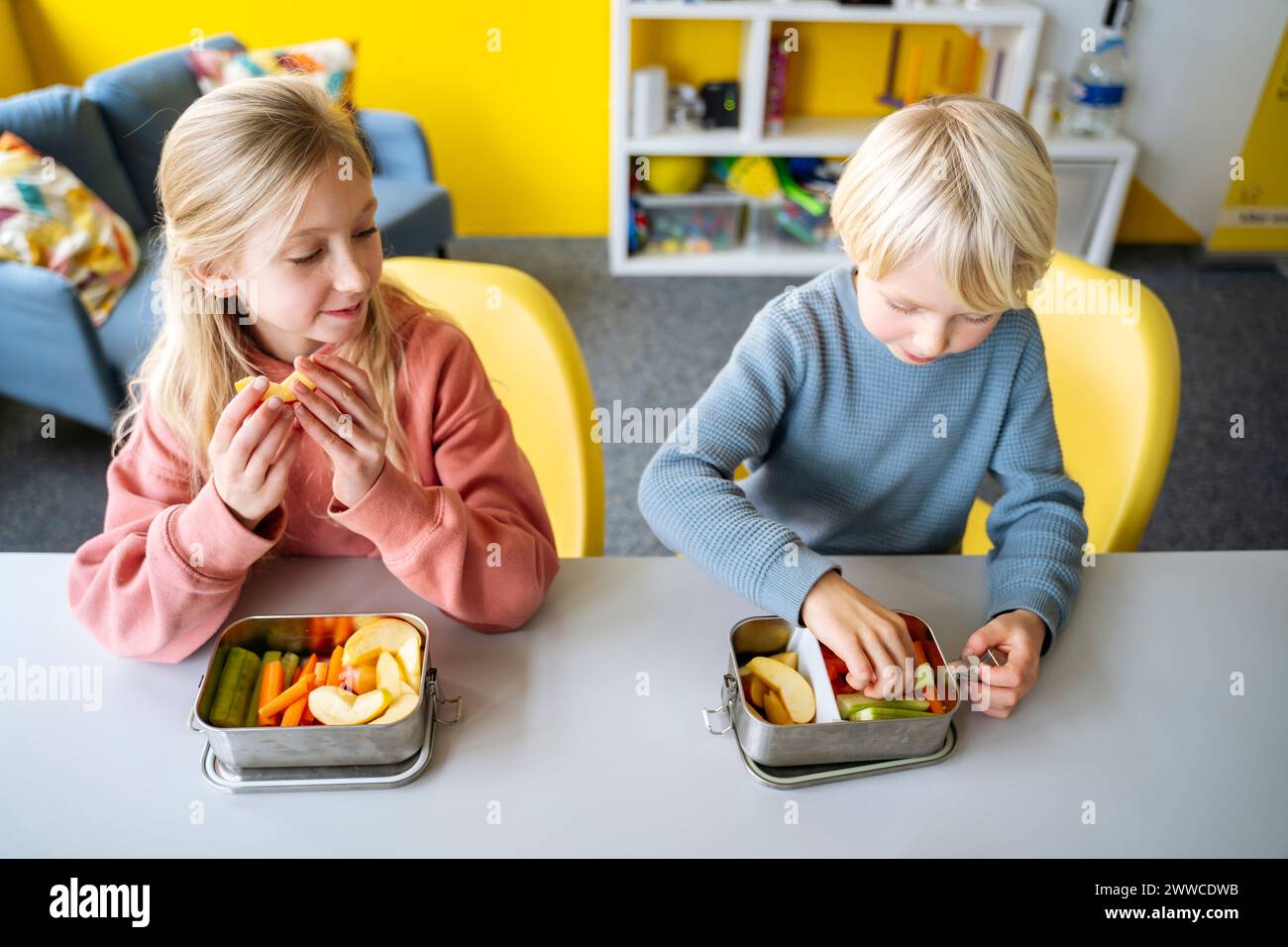 Blond boy and girl eating fruits and vegetables in lunch box at desk Stock Photo