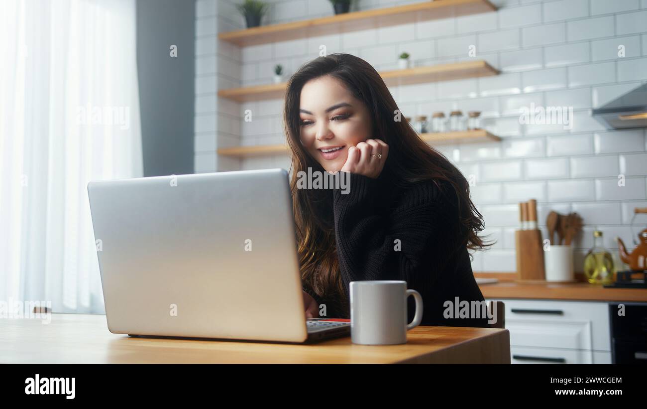 Cozy asian woman sitting in the kitchen using laptop computer surfing ...