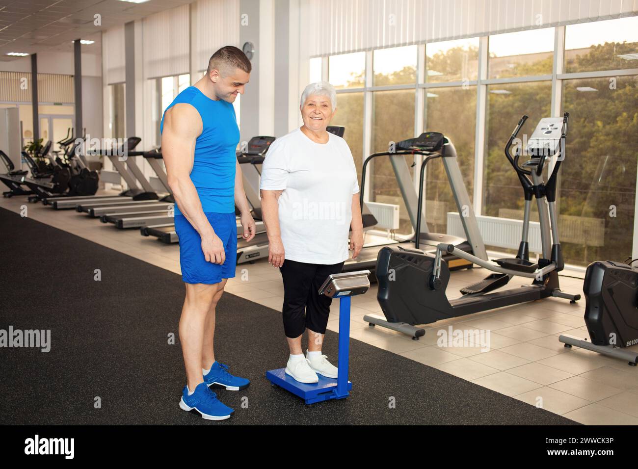 Pensioner stands on the scales, measures weight in the gym. Weight loss after training, physical activity, senior concept. happy, smiling, thin Stock Photo