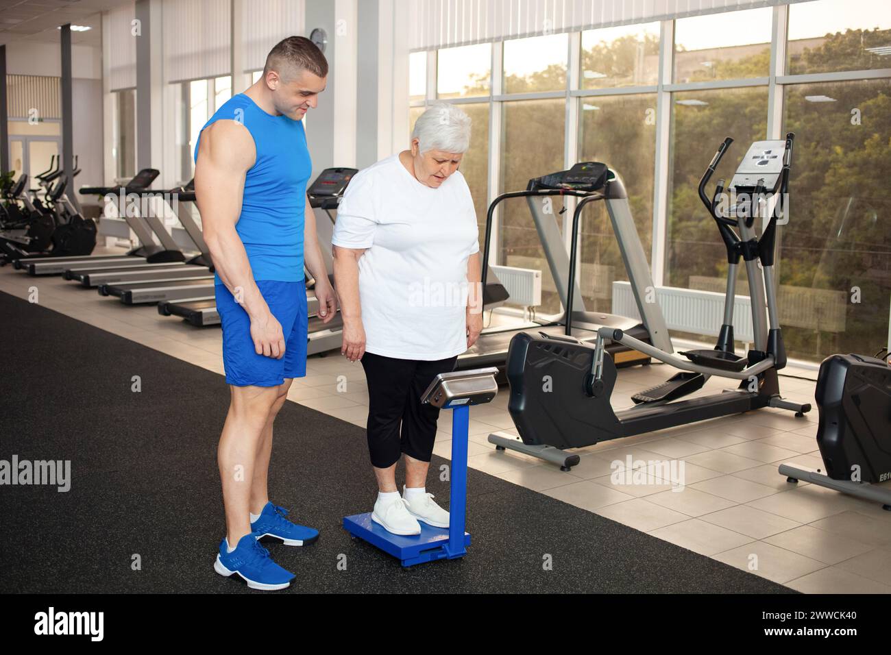 Pensioner stands on the scales, measures weight in the gym. Weight loss after training, physical activity, senior concept. sad, it doesn't work, tired Stock Photo