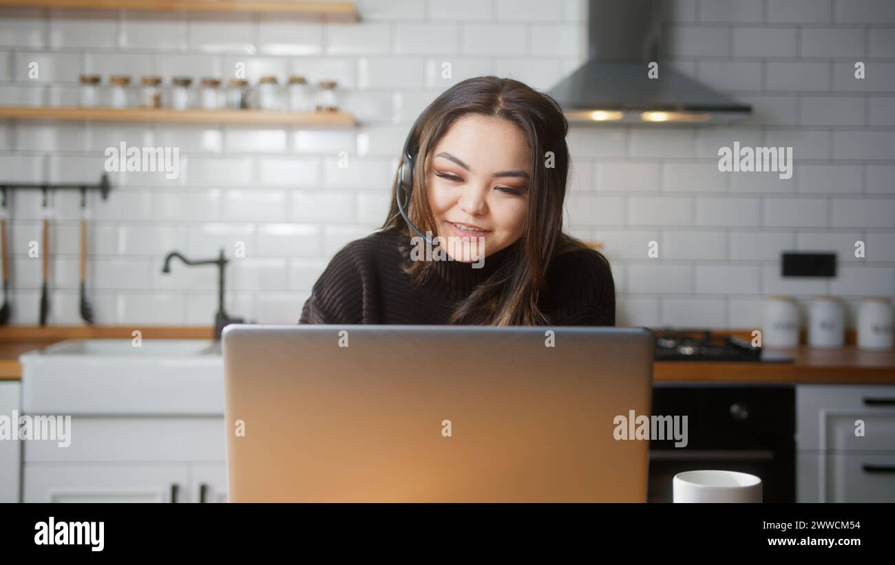 Young asian girl wear wireless headset having video conference call on laptop computer talk by webcam in online chat in kitchen at home. Customer supp Stock Photo